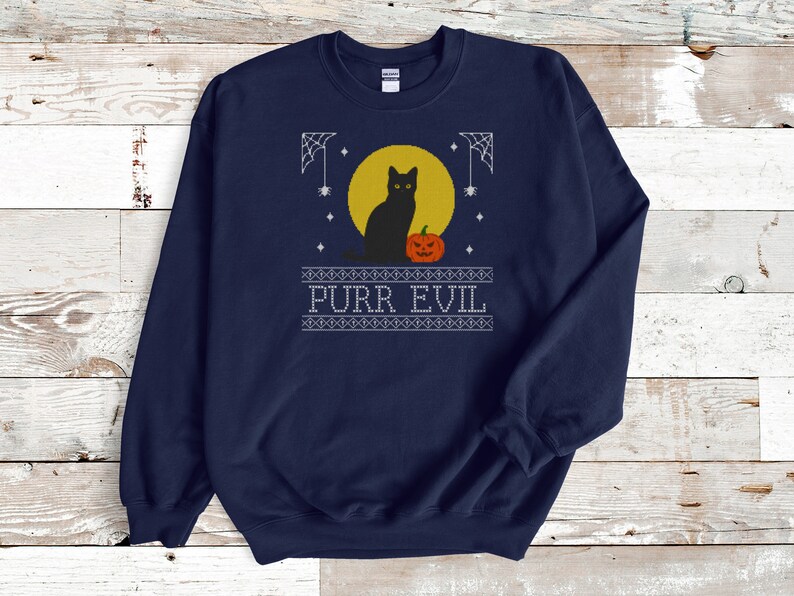 May include: A navy blue crewneck sweatshirt featuring a graphic print of a black cat sitting in front of a yellow full moon next to an orange jack-o'-lantern. The design includes white spiderwebs, stars, and the text PURR EVIL in a block font.