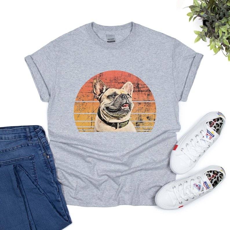 Graphic t-shirt featuring a French bulldog portrait with the name in bold, white, distressed font below. The dog image is placed inside a vintage-style, sunset-colored circle, blending shades of orange, yellow, and red for a retro aesthetic.