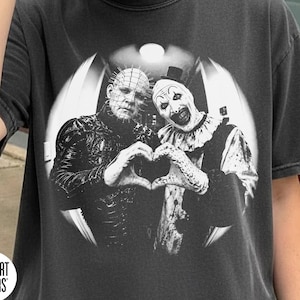 May include: A charcoal gray short-sleeve t-shirt featuring a circular black and white graphic of Pinhead and Art the Clown forming a heart shape with their hands. The shirt includes a Comfort Colors logo and the text Pepper Color.
