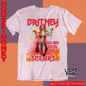 May include: Light pink t-shirt featuring a graphic of Britney Spears with the text "BRITNEY SPEARS" in red. The design includes an image of Britney holding a snake, with the words "AND NOW WATCH ME" in white. The shirt is labeled "LIGHT PINK".