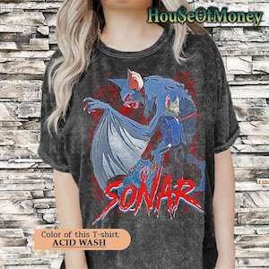 Puede incluir: Camiseta con lavado ácido con una criatura azul con forma de murciélago con detalles en rojo y la palabra "SONAR" en rojo. La camiseta también tiene una criatura verde con un traje azul. El color de la camiseta es lavado ácido.