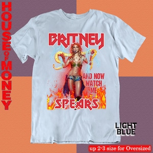 May include: Light blue t-shirt featuring a graphic of Britney Spears with the text "BRITNEY SPEARS" in red. The graphic includes the text "AND NOW WATCH ME" and a flame design. The shirt is labeled "LIGHT BLUE".