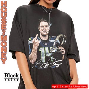 May include: Black oversized t-shirt featuring a graphic of a football player holding a trophy and smoking a cigar, with the number 14 and a signature. The shirt has the text "HOUSE OF MONEY" and "Black color".