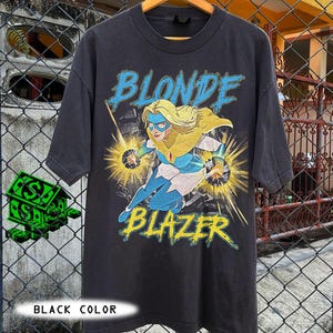 May include: Black t-shirt featuring a graphic of a blonde superhero with the words "BLONDE BLAZER" in blue and yellow. The superhero is depicted in a dynamic pose with energy bursts. The shirt is hanging on a wooden hanger.