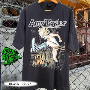 Może przedstawiać: Czarny t-shirt z grafiką z napisem "Amy Taylor" i ilustracją osoby w białym stroju i butach. Koszulka ma również napis "AMYL AND THE SNIFTENS". Koszulka jest w rozmiarze oversize.