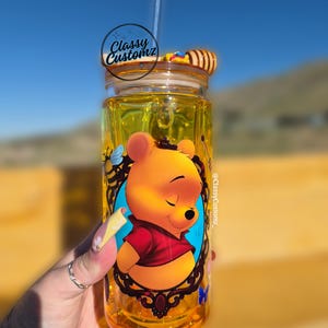 May include: A clear glass jar with a Winnie the Pooh design, filled with yellow liquid. The jar has a lid, a straw, and a decorative honey dipper. The jar is labeled "Classy Customz".