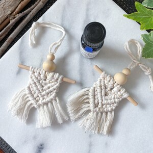 May include: Two white macrame ornaments with wooden beads and fringe. The ornaments are hanging from a string. A small bottle of essential oil is in the background.