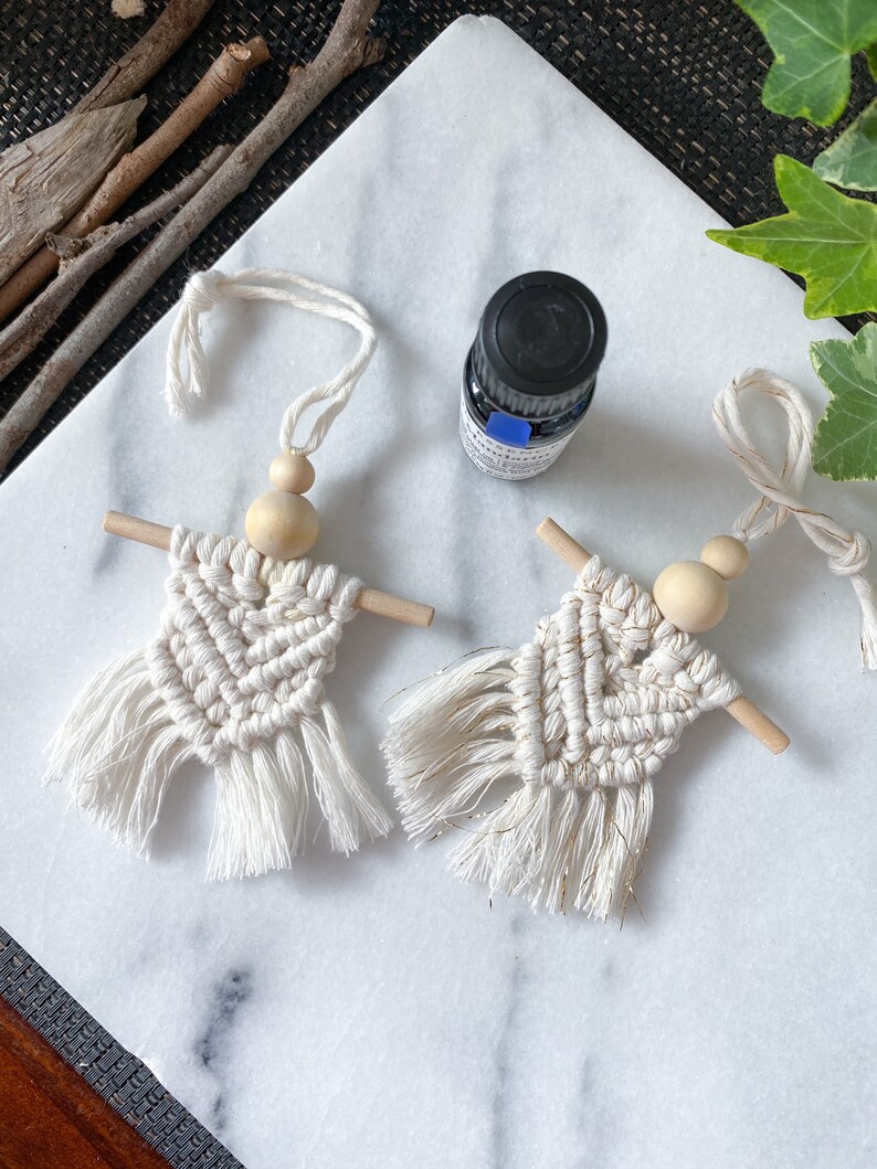 K&ouml;nnte beinhalten: Zwei wei&szlig;e Makramee-Anh&auml;nger mit Holzperlen und Fransen. Die Anh&auml;nger h&auml;ngen an einer Schnur. Eine kleine Flasche &auml;therisches &Ouml;l befindet sich im Hintergrund.