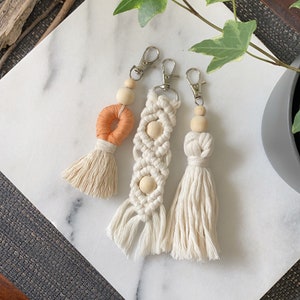 May include: Three handmade macrame keychains with wooden beads and tassels. The keychains are in shades of white and orange.