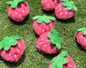 May include: Eight pink fuzzy strawberry-shaped ornaments with green felt leaves. The strawberries are arranged on a green grassy surface.