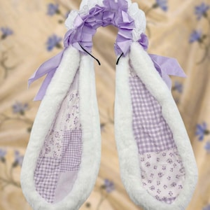 May include: A headband featuring two long, plush white faux fur ears with a patchwork interior pattern of lavender gingham and floral prints. The top of the headband is decorated with ruffled lavender satin fabric and white lace trim.