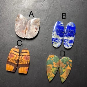 May include: Four pairs of polished gemstones are displayed. The gemstones include a pair of agate stones with brown and white patterns, two lapis lazuli stones with blue and white marbling, two stones with orange and brown patterns, and two green and orange patterned stones.