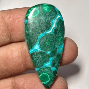 May include: A polished, teardrop-shaped gemstone. The stone's deep green base is patterned with vibrant turquoise blue veins. The gemstone is held in a hand, highlighting its natural colours and unique patterns.
