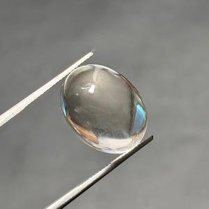May include: A clear, oval gemstone held by silver tweezers. The gem has a smooth, polished surface with subtle internal reflections and a slight iridescent sheen. The background is a muted grey.