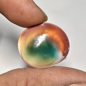 May include: A colorful, round glass marble with a swirl of orange, green, and white. The marble is held between two fingers, showcasing its polished surface and vibrant hues. The marble's design is reminiscent of a miniature planet.