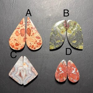 May include: Four pairs of polished gemstone cabochons. Pair A is teardrop-shaped with orange and red spots. Pair B is a semi-circle shape with green and brown patterns. Pair C is a triangle shape with gray and red bands. Pair D is teardrop-shaped with red and white spots.