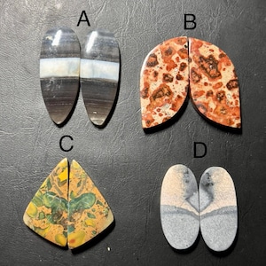 May include: Four pairs of polished gemstone cabochons. Pair A is dark gray with a white band. Pair B is red and tan with a speckled pattern. Pair C is yellow, green, and brown with a spotted design. Pair D is gray and tan with a landscape design.