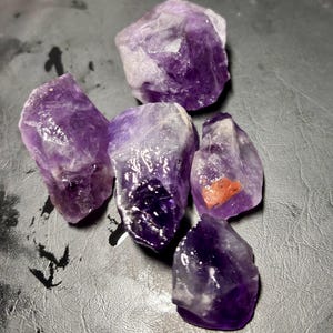 Könnte beinhalten: Eine Sammlung roher Amethystkristalle. Die Kristalle variieren in Größe und Form und zeigen eine Reihe von lilafarbenen Farbtönen, von hellem Lavendel bis zu tiefem Violett. Einige Kristalle haben einen rötlich-orangen Einschluss.