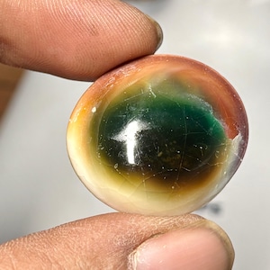 Puede incluir: Una piedra preciosa pulida y redonda con un llamativo degradado de color. La piedra pasa de una base blanco crema a tonos de amarillo, naranja y rojo, culminando en un centro verde oscuro. La superficie tiene grietas visibles.