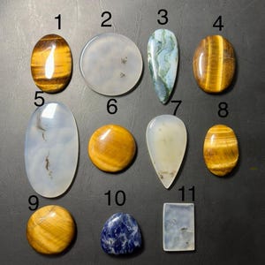 May include: A collection of eleven polished gemstones in various shapes and colors. The stones include tiger's eye, agate, and sodalite. The gemstones are oval, round, and teardrop-shaped, with a range of colors from golden brown to blue and white.