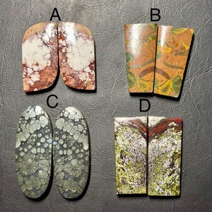 May include: Four pairs of polished gemstones are displayed. The first pair is a mix of white, brown, and red. The second pair is brown with green and yellow patterns. The third pair is oval-shaped with a gray, textured surface. The fourth pair is green and red.