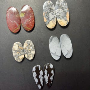 May include: A collection of six pairs of polished gemstone cabochons in various colors and patterns. The stones include red, gray, beige, and black, each with unique veining and textures. The shapes are oval and teardrop.