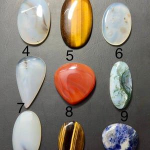 May include: Nine polished gemstones of various shapes and colors. The gemstones include oval, teardrop, and heart shapes. Colors include white, brown, red, green, and blue. The gemstones are arranged on a dark gray surface.