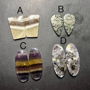 May include: Four pairs of polished gemstones are displayed. The gemstones are in various shapes and colors, including tan and brown, silver and gray, purple and yellow, and gray and purple. The gemstones are arranged on a dark surface.