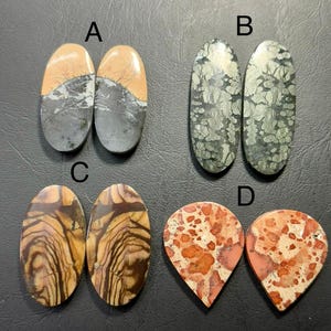May include: Four pairs of polished gemstones are displayed. Pair A features oval stones with tan, gray, and black patterns. Pair B showcases oval stones with a green, gray, and white mottled design. Pair C has oval stones with brown and tan wood grain patterns. Pair D has teardrop-shaped stones with red and orange spots.