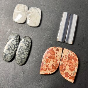 May include: A collection of polished gemstone cabochons in various shapes and colors. The stones include white and gray, gray and black, and red and tan patterns. The cabochons are oval, rectangular, and leaf-shaped.