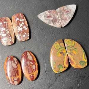 May include: A collection of polished gemstones in various shapes and colors. The stones include oval and rectangular shapes with patterns in shades of brown, red, white, and yellow. The gemstones are displayed on a dark surface.