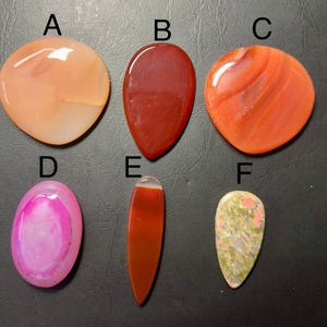 May include: Six polished gemstones in various shapes and colors. The stones include shades of orange, red, pink, and green. The gemstones are displayed on a dark surface.
