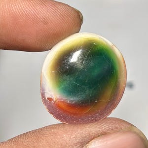 Puede incluir: Una gema redonda y colorida con una mezcla de colores que incluyen verde, amarillo, naranja y morado. La piedra tiene una apariencia agrietada y se sostiene entre dos dedos. La gema es probablemente un artículo decorativo o una joya.