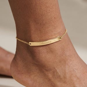 Peut inclure: Un bracelet de cheville doré avec un pendentif rectangulaire portant le nom "Alexandra" gravé. Le bracelet est attaché à une chaîne.