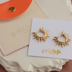 Puede incluir: Un par de pendientes dorados con forma de sol, expuestos sobre una tarjeta blanca con la marca "goldfolk LONDON". Una funda de cuero naranja con un broche dorado es visible junto a los pendientes. Los pendientes están sobre una superficie blanca texturizada.
