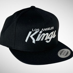 May include: Black baseball cap with a white embroidered logo that says "Los Angeles Kings". The cap has a black adjustable strap with a silver snap closure.