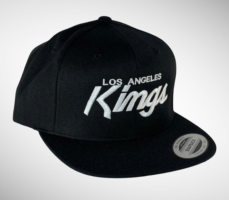 May include: Black baseball cap with a white embroidered logo that says "Los Angeles Kings". The cap has a black adjustable strap with a silver snap closure.
