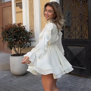 May include: A white long-sleeved mini dress featuring a high neckline, a cinched waist, and multiple layers of ruffles along the sleeves and the flared skirt. The garment is constructed with a solid white fabric and includes a tie detail at the neck.