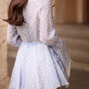May include: A pale blue dress featuring a white polka dot design. The dress has long sleeves, a gathered waist, and a flared skirt. The fabric looks lightweight and slightly transparent, with a soft drape.