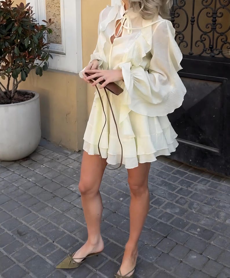 May include: A pale yellow mini dress featuring long, voluminous sleeves with ruffled cuffs, a tiered ruffled skirt, and a high neckline with a tie closure. The dress is paired with olive green pointed-toe mules and a small brown leather crossbody bag.