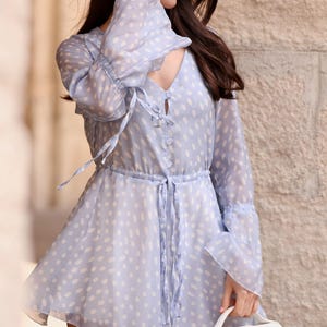 May include: A pale blue dress with white polka dots. The dress has long, flared sleeves, a V-neck, and a tie-waist. The outfit is completed with a white handbag.