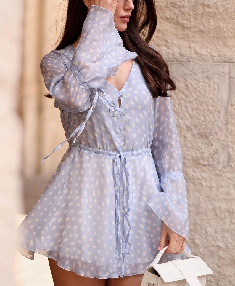 May include: A pale blue dress with white polka dots. The dress has long, flared sleeves, a V-neck, and a tie-waist. The outfit is completed with a white handbag.