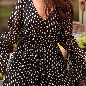May include: Black romper with white polka dots. The V-neck romper features long sleeves with flared cuffs and a cinched waist. The fabric appears to be a lightweight, semi-sheer material. The romper has a button-down front.