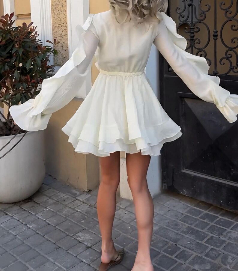 May include: A cream-colored mini dress featuring long sleeves with ruffled detailing, a cinched waist, and a multi-layered flared skirt. The garment is constructed from a lightweight, semi-sheer fabric with a soft, flowing silhouette throughout.