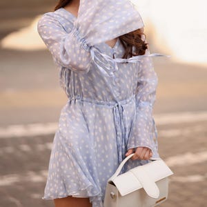 May include: A pale blue dress with white spots, long sleeves with flared cuffs, and a drawstring waist. The dress is worn with a white handbag with a top handle. The dress has a V-neck and a tie at the waist. The dress is knee-length.