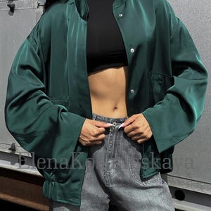 Pode incluir: Casaco bomber de cetim verde escuro com gola redonda e fechos de pressão. O casaco tem dois bolsos frontais e é combinado com jeans cinza. O texto "ElenaKoshskaya" é visível na imagem.