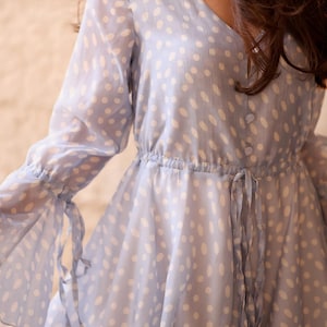 May include: A pale blue dress with a V-neck and long sleeves. The dress has a white polka dot pattern and a cinched waist with a tie. The sleeves have ties at the wrists, and the skirt is flowing.