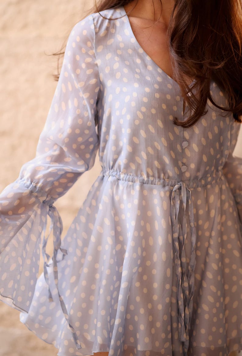 May include: A pale blue dress with a V-neck and long sleeves. The dress has a white polka dot pattern and a cinched waist with a tie. The sleeves have ties at the wrists, and the skirt is flowing.