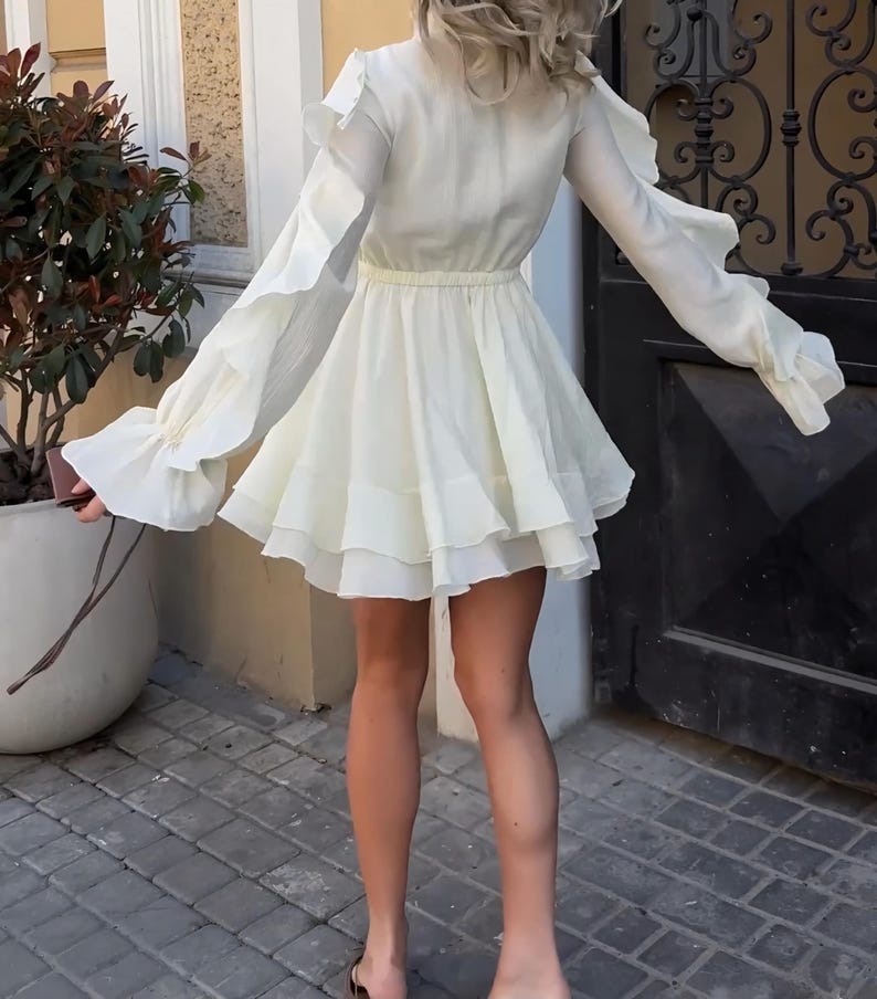 May include: A cream-colored long-sleeve mini dress featuring a high neckline, a cinched waist, and layered ruffle detailing along the sleeves and the skirt hem. The garment is constructed from a lightweight, semi-sheer fabric with a flowing silhouette.