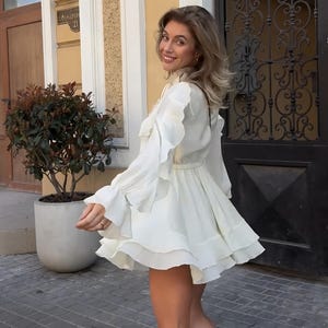 May include: A white long-sleeved mini dress featuring a high neckline, a cinched waist, and multiple layers of ruffles along the sleeves and the flared skirt. The garment is constructed with a solid white fabric and includes a tie detail at the neck.